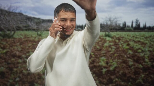 Man on phone smiling, raising right hand palm to camera in field wearing cream sweater; greeting happiness.