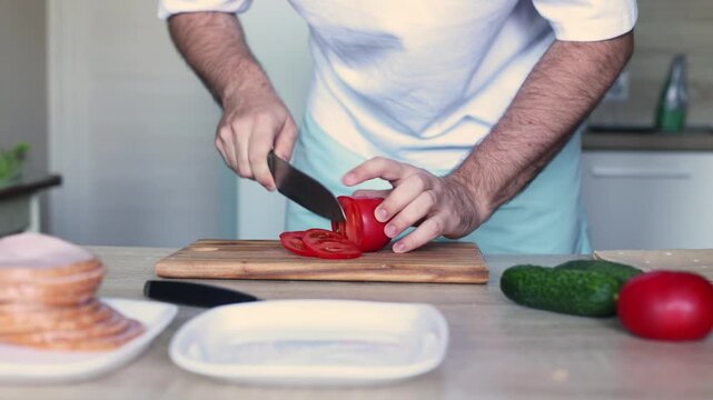 A man slices a red tomato on a wooden board in a bright kitchen, fresh cucumbers, bread, and plates nearby, evoking mindful meal prep, home cooking, and wholesome daily ritual.