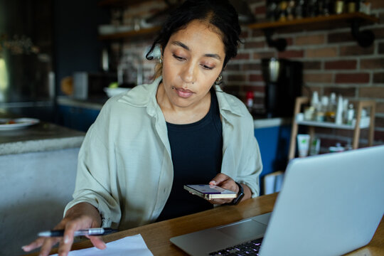 Young woman working from home kitchen with laptop and smartphone