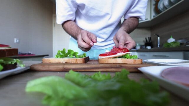 Hands layer lettuce, tomato, ham, and cheese onto bread slices on a wooden board, capturing mindful sandwich assembly, nourishment, and everyday care.