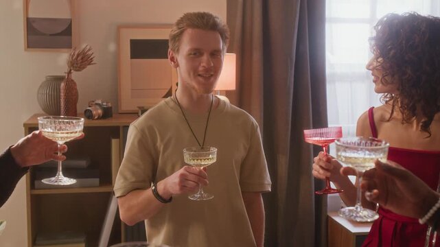 Tilt up view of smiling young man proposing toast looking at his friends, then they clinking glasses together during home celebration