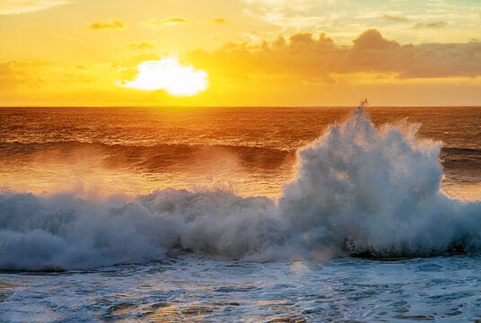 Dramatic ocean waves crashing under warm golden sunset light with glowing clouds and dynamic sea spray.