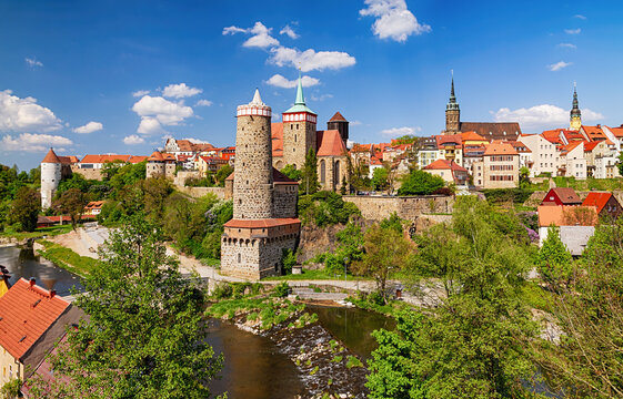 Panoramic view of Bautzen&rsquo;s historic old town with medieval stone towers, red-tiled roofs, and the Spree River winding through lush greenery under a bright blue sky.