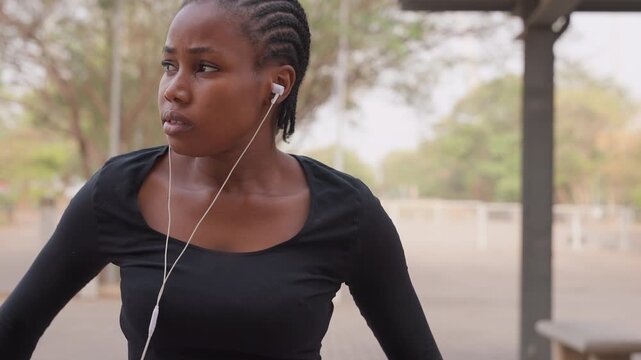 black woman sitting under shelter listening to music through earbuds, cornrow braids, casual black top, empty street and park trees in background, pensive gaze, side glances, late afternoon light,