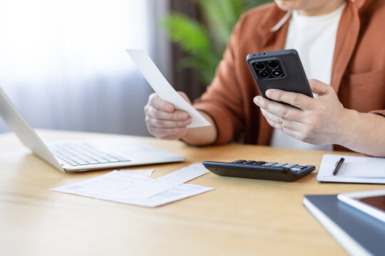 Person calculating financial budget, analyzing invoices with a mobile application, managing personal finance, and planning household expenses on a wooden desk