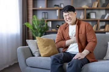 Fotobehang Onderzeeër Asian man experiencing a sudden heart attack or angina while sitting on a living room sofa, clutching his chest with an expression of intense discomfort and medical emergency  © Liubomir