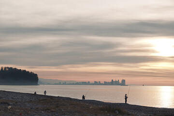 Beach at sunset with calm water and silhouettes of people fishing along the shore, distant city...