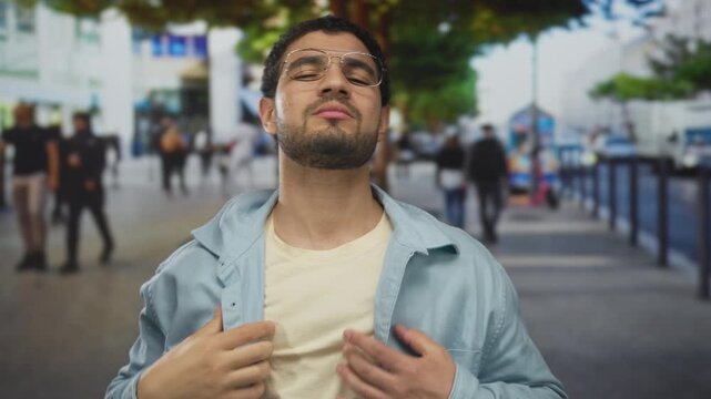 Man with glasses and beard adjusting his jacket and touching his chest on a busy city street; confidence.
