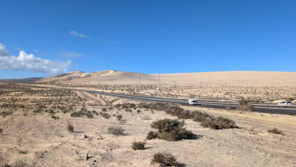 Asphalt road through arid landscape with sand, rocks, and dry bushes in Fuerteventura, Spain. 