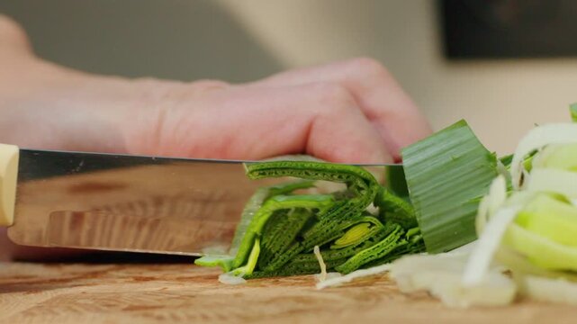 A hand cuts leeks on a wooden board in a kitchen. The green parts of the leeks are neatly arranged