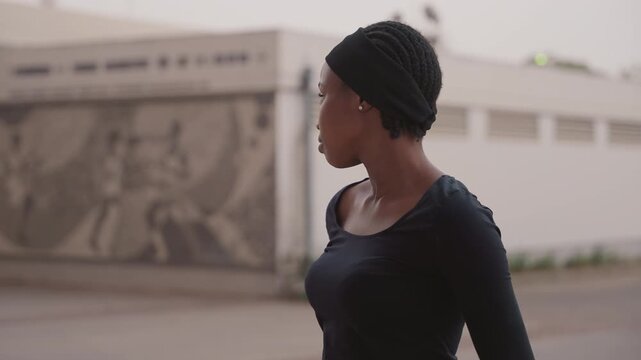 black woman walking past mural at dusk, determined stride and alert profile capturing motion and confidence, streetwear outfit with scarf, energetic posture and timely turn toward sidewalk, commuter