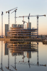 High rise building construction by the waterfront at sunrise, multiple tower cranes reflected in calm water as concrete floors rise into the city skyline   © Nannie