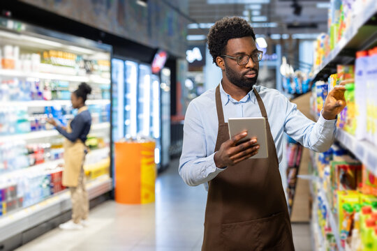 Retail worker holding a digital tablet while performing inventory management and restocking shelves with products at a grocery store, ensuring efficient store operations
