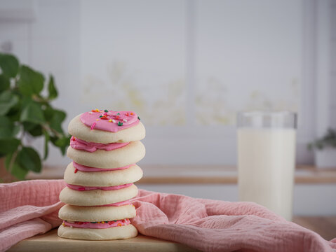 Yummy delicious Stack of pink frosted Spring Easter sugar cookies with tall glass of cold milk and cloth muslin fabric napkin on wood table counter with plant and white kitchen backdrop