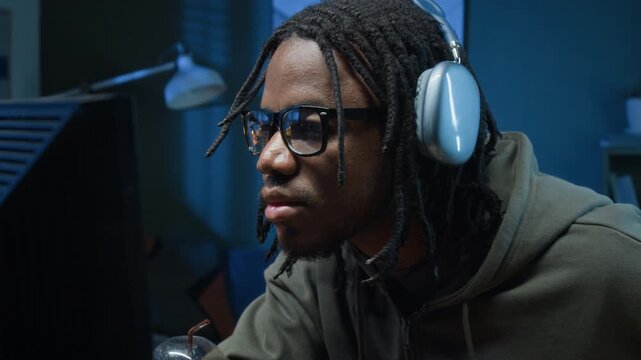 Focused Black male hacker wearing headphones and glasses typing code on computer keyboard reading screen while cracking passwords during late night in private workspace