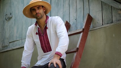 Young ukrainian man wearing an embroidered shirt looking into camera outdoor. Portrait of guy in straw hat against the background of his house. Concept of national identity and patriotism