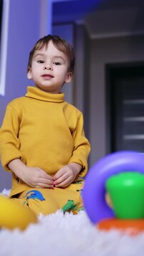 Funny boy in yellow clothes sits on the carpet. Kid plays with rings from toy pyramid. Low angle view. Vertical video