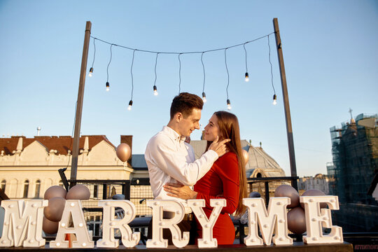 Couple enjoys special moment on rooftop with lights and sign asking for marriage