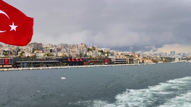 Cinematic View of Turkish Flag Waving on a Boat Trip Towards Eminonu with Galataport, Modern Buildings, and Dramatic Stormy Sky Over Istanbul Bosphorus
