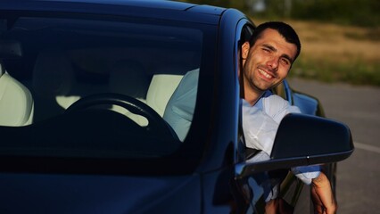 Young smiling entrepreneur in shirt leaning out of driver window at modern electric car and looking into camera. Portrait of handsome successful businessman in new electrical vehicle. Slow mo
