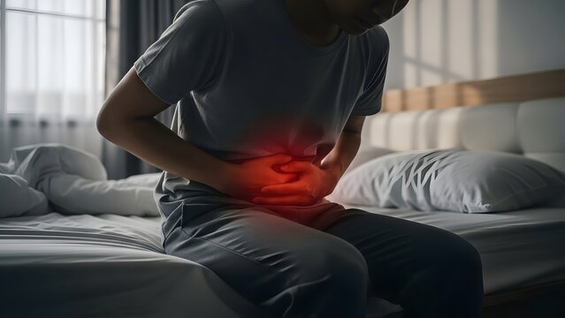 Person suffering from severe stomach pain, highlighted red, while sitting on bed. Abdominal discomfort and illness concept.