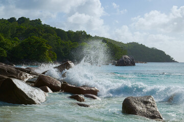 Powerful waves crash against granite rocks on a tropical Seychelles beach, surrounded by lush green hills and clear blue water.
