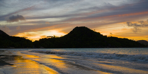 A peaceful Seychelles beach at sunset, with gentle waves reflecting golden light and a lush island silhouette on the horizon, capturing a serene vacation moment in paradise.
