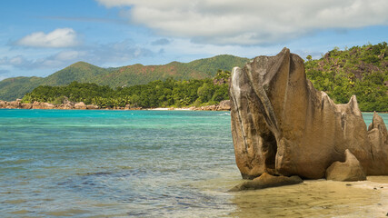 A tropical beach in the Seychelles with clear turquoise water, granite rocks, and lush green hills, capturing a peaceful vacation travel moment.
