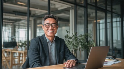 Fototapeta premium Professional adult Asian man smiling at his desk with a laptop in a bright office setting