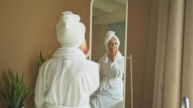 Mature adult old woman looking at mirror applying nourishing anti age face cream doing daily beauty routine. Senior woman with white mask creme on skin gets facial treatment Skin care aging skincare
