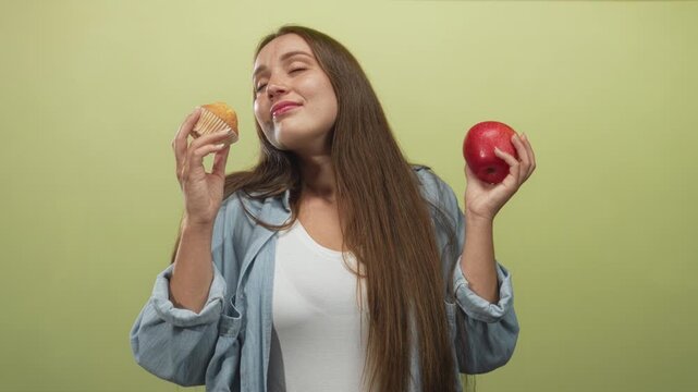 Woman holding a red apple and a cupcake in a light green studio, glancing at the apple while comparing choices; choice indecision.