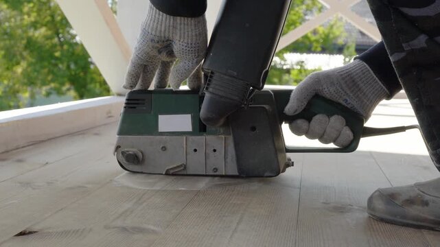 Carpenter smooths wooden floor with electric sander outdoors on sunny day