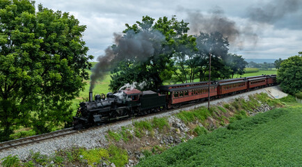 A steam locomotive pulls several carriages along the railway surrounded by lush greenery. Smoke rises from the engine as it moves past the trees on an overcast day.