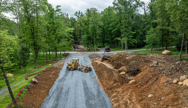 Workers are using machinery to build a road in a forested area. The ground is being leveled, and gravel is laid in the foreground. Trees surround the site, and a path is visible in the distance.