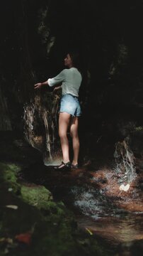 Female tourist is carefully walking on rocks in a cave, behind a small waterfall, discovering the beauty of tropical nature