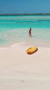 Young woman standing in shallow crystal clear water on a pristine white sand beach with a yellow kayak nearby, enjoying the turquoise lagoon and the tropical island in the background