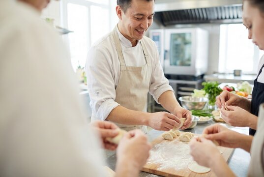 A smiling Asian head chef teaches an interactive cooking class, showing a group of students how to form dumplings in a professional kitchen setting.