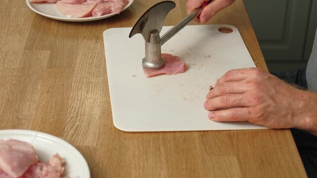 Person using meat tenderizer on raw chicken. Chef preparing pork loin slice with meat mallet on cutting board. Man tenderizing fillet. Cooking cutlets pounding poultry breast close-up. Culinary dish.