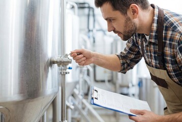 A brewery worker inspects a stainless steel fermentation tank, checking a valve and taking notes on a clipboard.