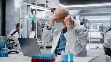 Stressed elderly researcher wearing surgical mask in laboratory using laptop software, driving treatment discovery. Anxious senior man overwhelmed wile working with bioinformatics app in lab, camera B