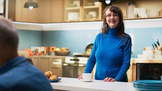 Senior wife standing in bright kitchen at home enjoying coffee cup and peaceful relaxation. Aged woman pensioner conveys positive retired life, wellbeing and joyful household moments. Camera B.