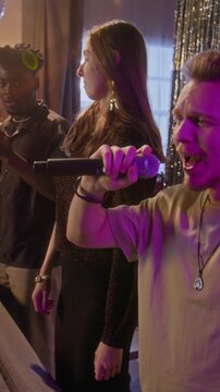 Vertical view of excited Caucasian guy in beige t-shirt energetically flailing his arm and performing song into microphone during karaoke night with friends at home