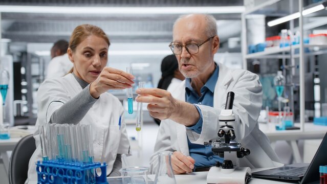 Lab team leader teaching intern to analyze liquid polymer properties in test tube. Senior scientist mentoring trainee to test new synthetic material viscosity in laboratory, camera B