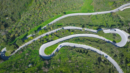 Obraz premium AERIAL TOP DOWN: Narrow mountain road carving tight hairpin turns through hillside covered with lush green Mediterranean greenery. Vehicles overcome serpentines through countryside in western Sicily.