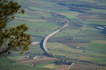 Obraz premium Curving highway viaduct crossing farmland in western part of Sicily. Soft, blurred branches in the foreground add depth and natural framing, contrasting with the clean geometry of the winding road.