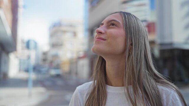 Young woman looking up on city street, face and bare neck visible, wearing white t shirt and gold hoop earring while standing near buildings; serenity.