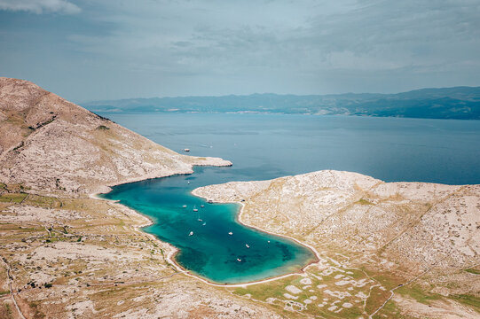 Aerial view of a turquoise bay cradled by sun-baked hills, where tiny boats dot the clear water under a vast sky, Baska, Primorje-Gorski Kotar County, Croatia.