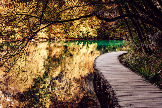 View of a wooden pathway winding along the still, turquoise waters reflecting vibrant autumn foliage, Plitvicka Jezera, Lika-Senj County, Croatia.