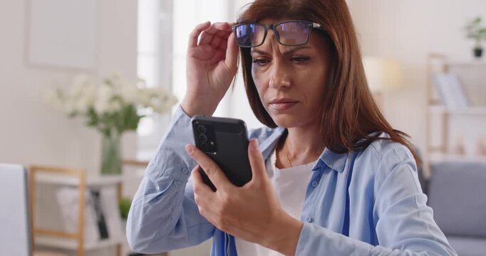 Woman with poor vision problem squinting and removing glasses to read blurry text on smartphone screen. Myopia, glaucoma and loss eyesight, digital eyestrain trouble of person working at home