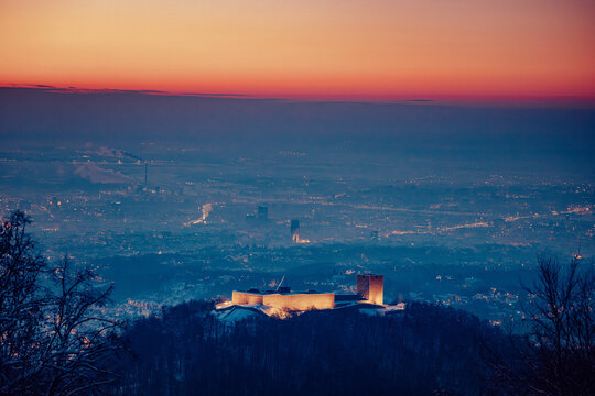 View of a medieval castle atop a hill bathed in the warm glow of twilight as the city lights flicker below against a serene, colorful sky, Sljeme, Croatia.